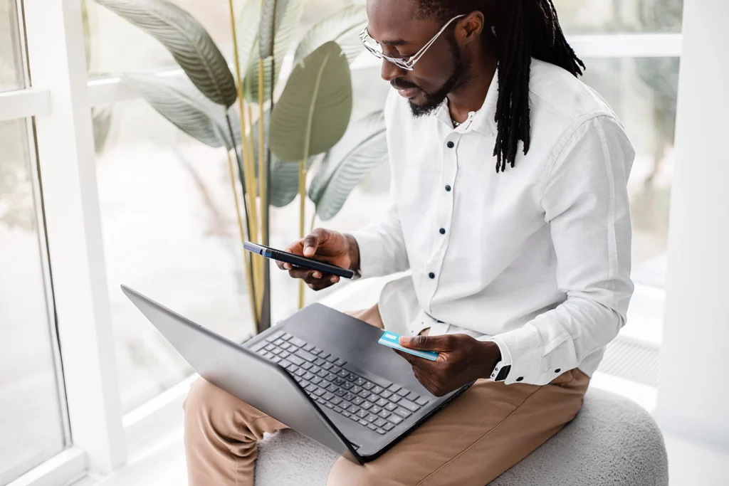 Country-Bank-man-sitting-on-white-chair-holding-phone-and-debit-card-with-laptop-on-his-lap