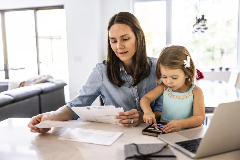 Country-Bank-mother-sitting-at-table-paying-bills-while-daughter-plays-with-phone