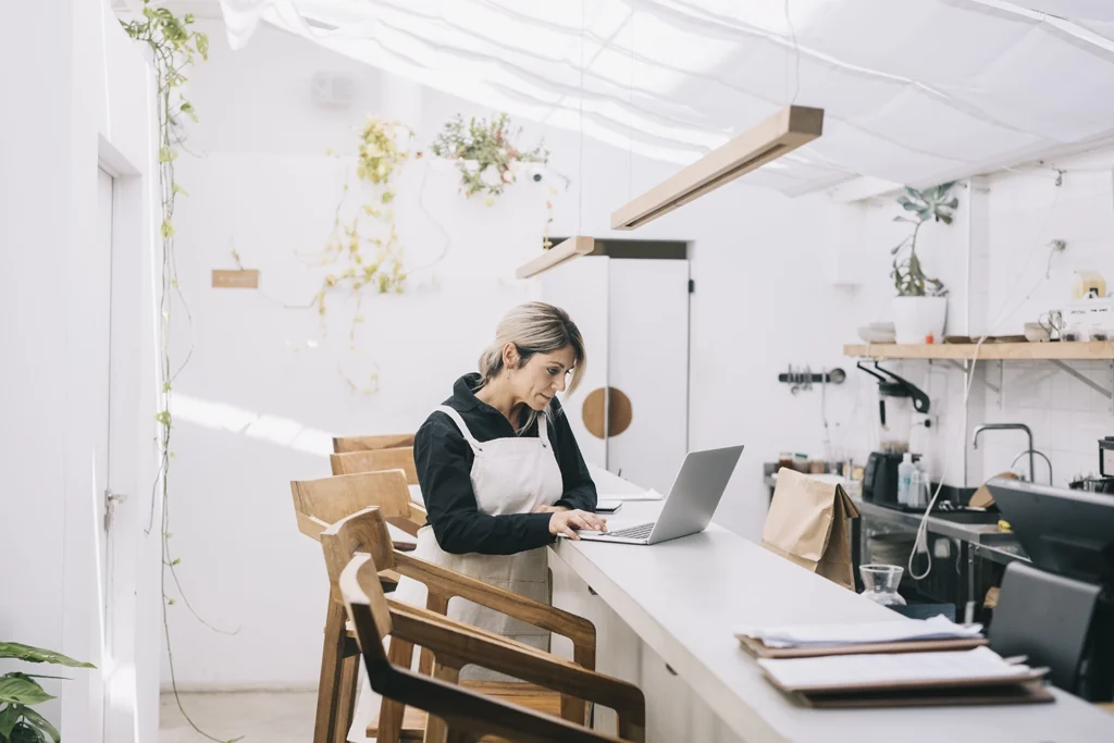 Country-Bank-woman-in-apron-sits-at-computer-at-bar-in-industrial-kitchen